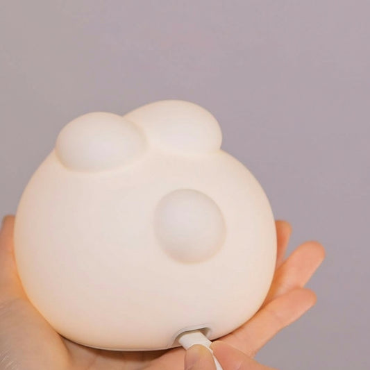 close-up shot of a person holding a soft, white, round night light that has small, rounded protrusions resembling ears and a face. A white charging cable is plugged into the bottom of the light.