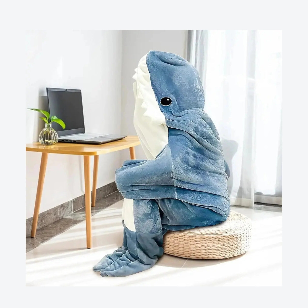 Person sitting on a chair covered with a blue shark-shaped blanket in a room with a laptop on a table.