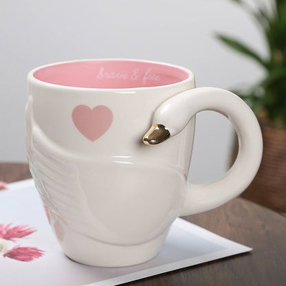 A white ceramic coffee mug with a pink and gold swan pattern and an ergonomic handle, displayed on a table with a plant in the background.