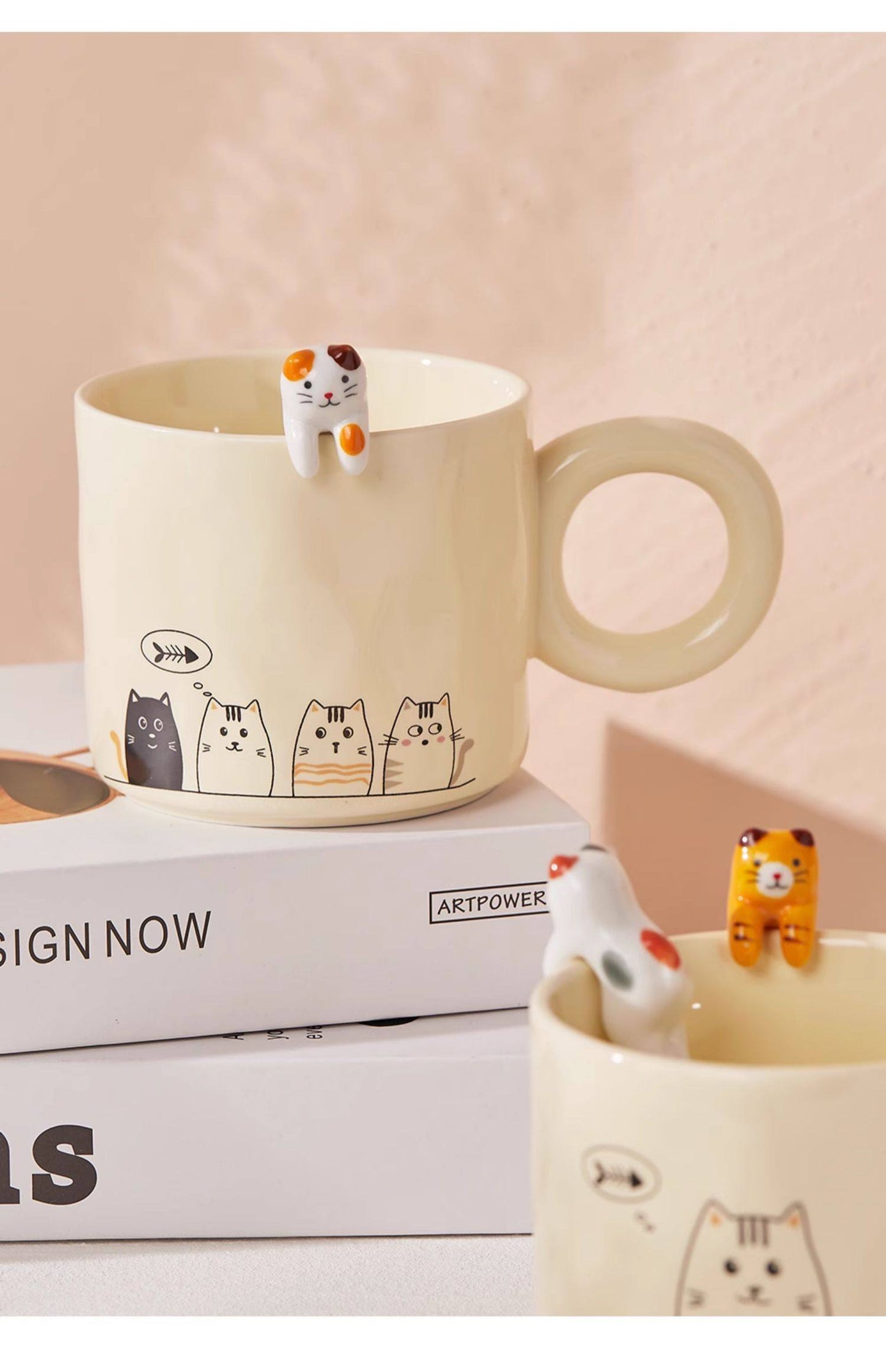Two ceramic mugs with cat-themed designs and small cat figurines on a light pink background.